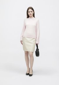 Žena oblečená v svetloružovom svetri a béžovej sukni, držiaca malú čiernu kabelku, stojaca s neutrálnym výrazom na bielom pozadí.