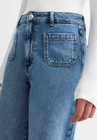 Pantalones vaqueros de mezclilla azul claro con cintura alta, diseño de cinco bolsillos, costuras visibles y textura suave. Cierre frontal con botón y cremallera.