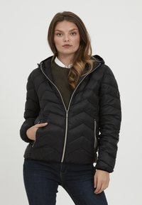 Doudoune noire avec un motif en chevrons, fermetures éclair grises, une capuche et des poches latérales ; portée sur un pull vert et une chemise blanche.