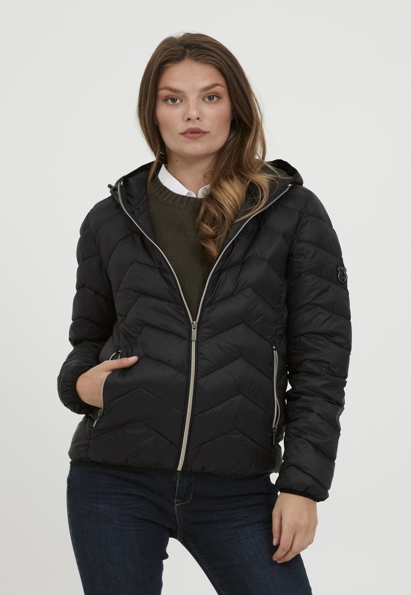 Doudoune noire avec un motif en chevrons, fermetures éclair grises, une capuche et des poches latérales ; portée sur un pull vert et une chemise blanche.