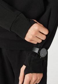 Sudadera negra con puños acanalados y un agujero para el pulgar. Combinada con un reloj negro que presenta un diseño minimalista con una correa texturizada oscura.