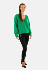 Maglione verde a scollo a V con vestibilità ampia e polsini a costine, abbinato a pantaloni neri attillati e scarpe con tacco nere.