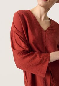 Rød, struktureret V-hals bluse med brede ærmer. Stoffet har en subtil glans og en let krøllet overflade. Har en central søm.