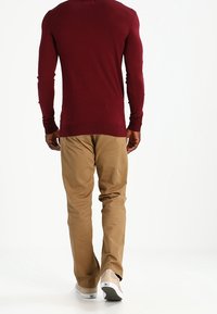 Bordeauxrode trui met lange mouwen en nauwsluitende mouwen, gecombineerd met beige chino's. De schoenen zijn lichtgrijs met een patroonaccent.