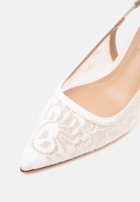 Λευκό παπούτσι slingback με μυτερή μύτη, με διακοσμητική δαντέλα με λουλούδια και μπεζ εσωτερική σόλα σε απλό φωτεινό φόντο.
