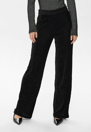 Schwarze weite Hose aus strukturiertem Stoff, mit hoher Taille und vertikalen Streifen, kombiniert mit spitzen schwarzen Schuhen.