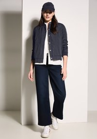 Marineblau bomberjacke über einem weißen Hemd, kombiniert mit weiten dunklen Jeans und weißen Sneakers. Das Model trägt außerdem eine marineblaue Mütze mit Text.