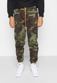 Kamouflaged mjukisbyxor i grön, brun och svart med elastiska muddars och en orange dragsko; har en liten orange logotyp på låret.