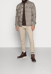 Chemise à carreaux marron et beige, chinos beiges et bottes en cuir marron foncé avec semelles blanches. Le modèle se tient contre un fond uni.