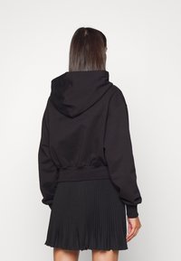 Černá zkrácená mikina s velkou kapucí, žebrovanými manžetami a pasem, v kombinaci s plisovanou černou sukní. Měkká textura, minimalistický design.