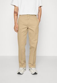 Man med beiga raka byxor, vita sneakers och en vit t-shirt, stående med händerna i fickorna mot en enfärgad bakgrund.