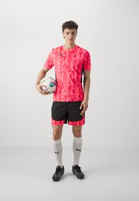 Camiseta deportiva gráfica de color rosa brillante con patrones geométricos, combinada con pantalones cortos negros con acentos rosa. Sostiene un balón de fútbol multicolor.
