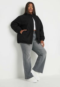 Czarna, teksturowana, polarowa kurtka z wysokim kołnierzem, zapięciem na guziki oraz ściągaczami na rękawach, w połączeniu z szarymi dżinsami w kształcie dzwonów i białymi sneakersami.