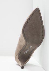 Beżowy zamszowy but na wysokim obcasie, z zaostrzonym noskiem, wąskim obcasem i subtelnymi przeszyciami. Rozmiar oznaczony jako 37. Widoczna nazwa marki.