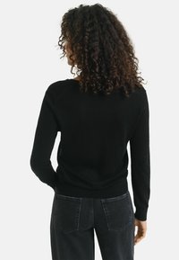 Schwarzer Pullover mit rundem Ausschnitt, langen Ärmeln und geripptem Saum. Getragen mit hoch taillierten schwarzen Jeans. Weiche Textur und figurbetonter Schnitt.