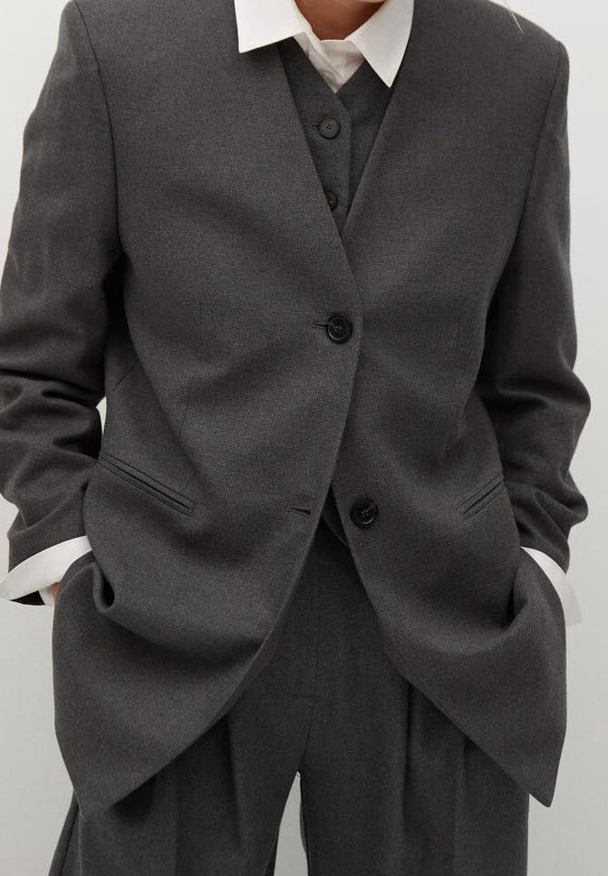 Personne portant un costume trois pièces gris foncé avec une chemise blanche, les mains dans les poches, debout devant un fond clair uni.