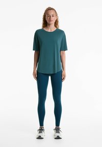 T-shirt teal à manches courtes avec un col rond associé à des leggings teal. Les baskets ont un design blanc et noir. Texture de tissu lisse.