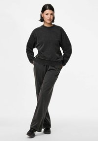 Jauna suaugusi moteris stovi su rankomis kišenėse, vilkinti juodą per didelį džemperį, derančias plačias kelnes ir juodus sportinius batelius.