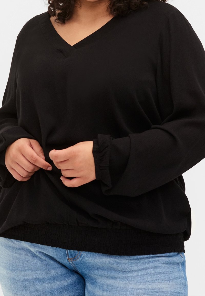 Haut noir à manches longues avec un col en V, un ourlet élastiqué et des poignets froncés. Le tissu semble doux avec une texture lisse. Associé à un jean bleu clair.