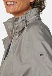 Beige wasserdichte Jacke mit hohem Kragen, Reißverschluss vorne, silbernen Akzenten und einem Logopatch auf dem linken Ärmel. Glattes, glänzendes Material.