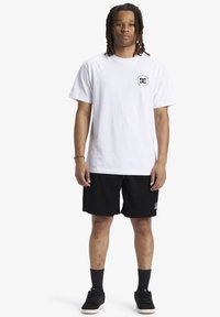 T-shirt bianco in cotone con un piccolo logo nero sul petto; abbinato a pantaloni corti atletici neri a trama e sneakers nere.