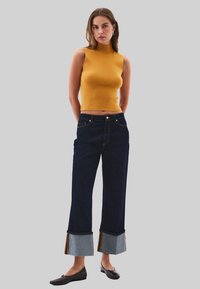Mosterdkleurige mouwloze top, donkerblauwe wijde broek met denim en grijze boorden, en zwarte flats. Strak zittende, gestructureerde stof met zichtbare naden.