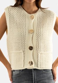 Veste en maille blanc cassé, avec un motif texturé, quatre gros boutons et des poches frontales. Sans manches avec une coupe décontractée.