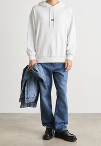 Sudadera blanca con bolsillos frontales, combinada con unos vaqueros de mezclilla azul sueltos. Una chaqueta de mezclilla está colgada de un brazo y lleva zapatos negros.