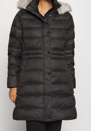Manteau d'hiver noir matelassé jusqu'aux genoux avec fermeture à boutons et capuche bordée de fourrure, porté par une personne les mains le long du corps.