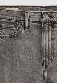 Gros plan sur un jean en denim gris montrant la poche avant, la passants de ceinture, un bouton en métal, et une petite étiquette en tissu indiquant "Quality Clothing".