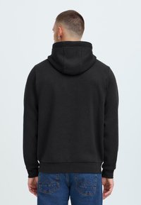 Svart hettegenser i myk stoff, med en stor hette og ribbede mansjetter, sammen med blå denimjeans med karakteristisk baklomme-stikking.