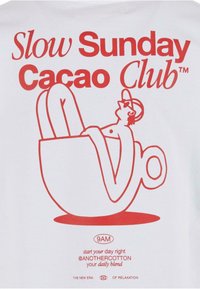 Hvit t-skjorte med røde grafikker av en figur som slapper av i en kakao-kopp med teksten: "Slow Sunday Cacao Club" og ytterligere merkevaredetaljer.