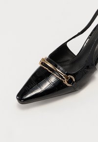 Chaussure slingback pointue en cuir verni noir avec texture crocodile et détail chaîne dorée sur l’orteil.