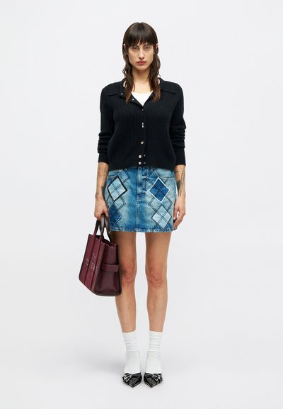 Fekete begombolt kardigánban, fehér toppal, kék gyémántmintás farmer szoknyában, fehér zokniban, fekete cipőben álló nő, aki bordó táskát tart.