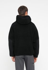 Veste en polaire noire avec un col haut, tissu texturé et coupe oversize. Présente des accents de panneaux noirs sur les manches.