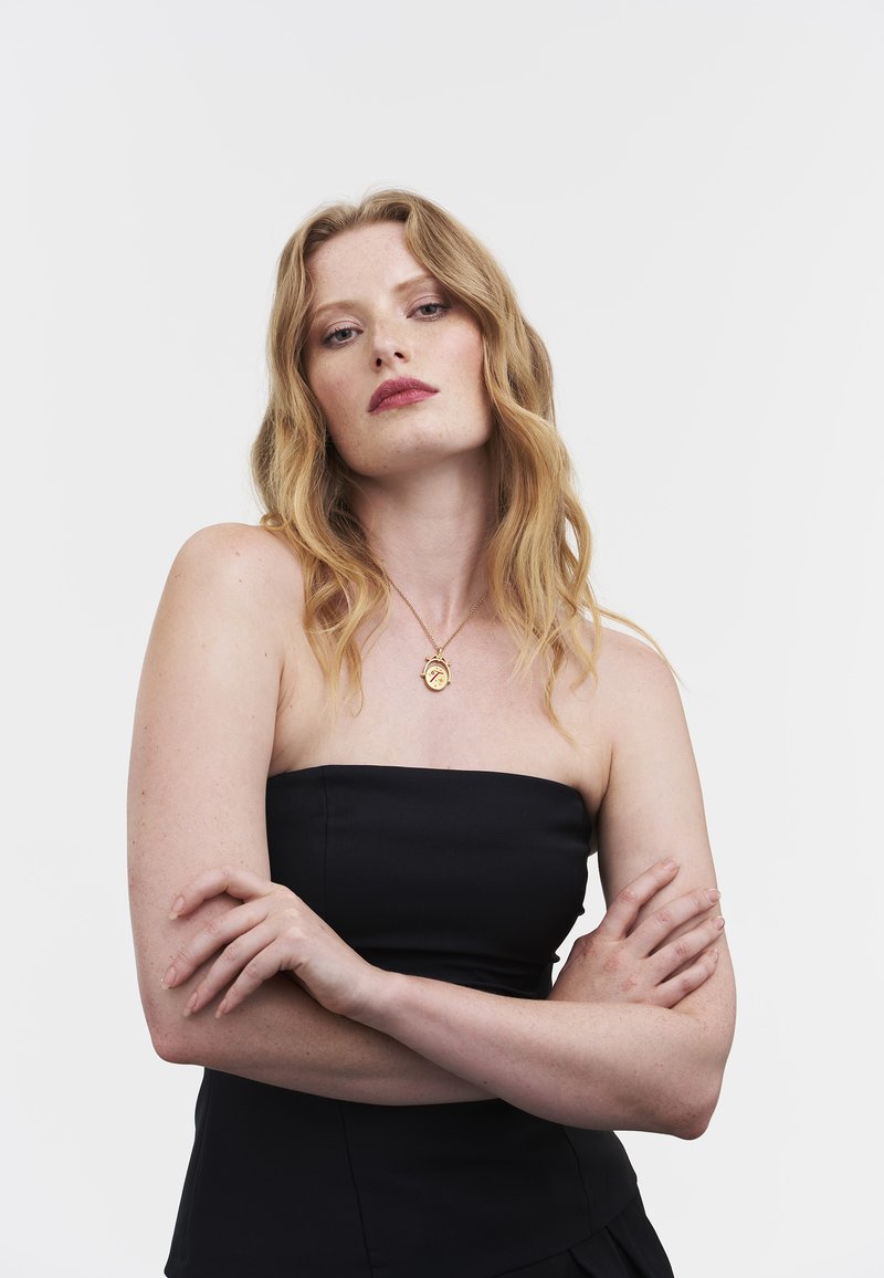 Gouden hangersketting gedragen door een model met lang, golvend haar. Ze draagt een zwarte, strapless top, met gekruiste armen, tegen een effen witte achtergrond.