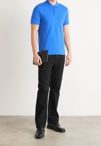 Modrá polo košile s límečkem a knoflíky, v kombinaci s černými kalhotami a černými botami. Jedinec drží v jedné ruce černý tablet.