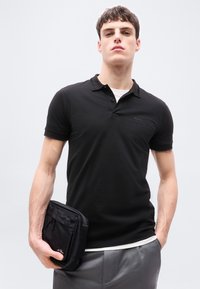 Νέος άνδρας με μαύρο κοντομάνικο πόλο και γκρι παντελόνι, κρατώντας μια μικρή μαύρη θήκη με φερμουάρ στο αριστερό του χέρι.