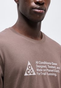 Κοντινό πλάνο ενός ατόμου που φορά ένα καφέ πουκάμισο με λευκό κείμενο: «Εξοπλισμός για όλες τις συνθήκες. Σχεδιασμένο, δοκιμασμένο και κατασκευασμένο στον πλανήτη Γη, για trail running.»