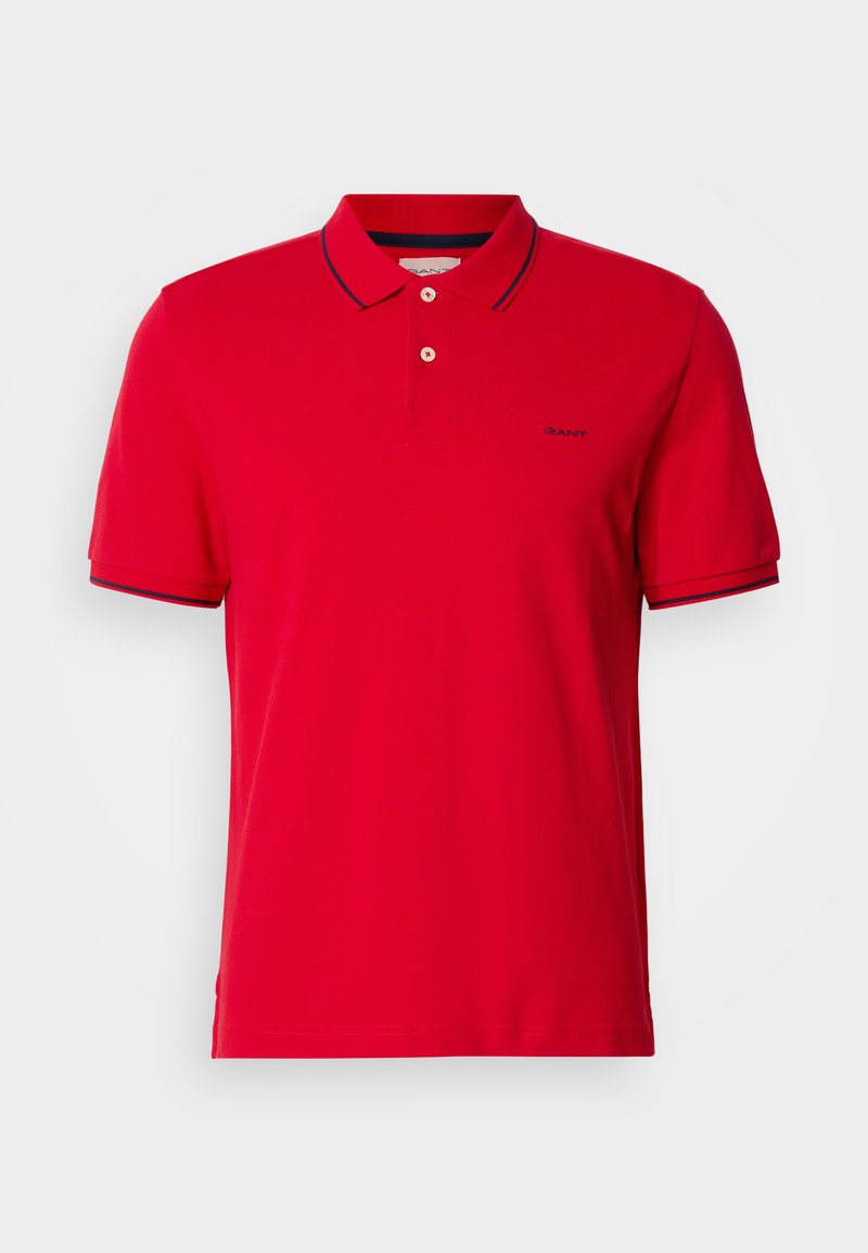 Polo-shirt rouge en coton, avec un col classique, une patte de boutonnage à deux boutons et des accents rayés bleu marine sur les manches et le col. Logo GANT présent.