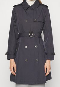 Mörkblå trenchcoat med dubbelknäppt design, stora knappar, justerbart bälte och axelroyaler. Har sidofickor och klassisk krage.