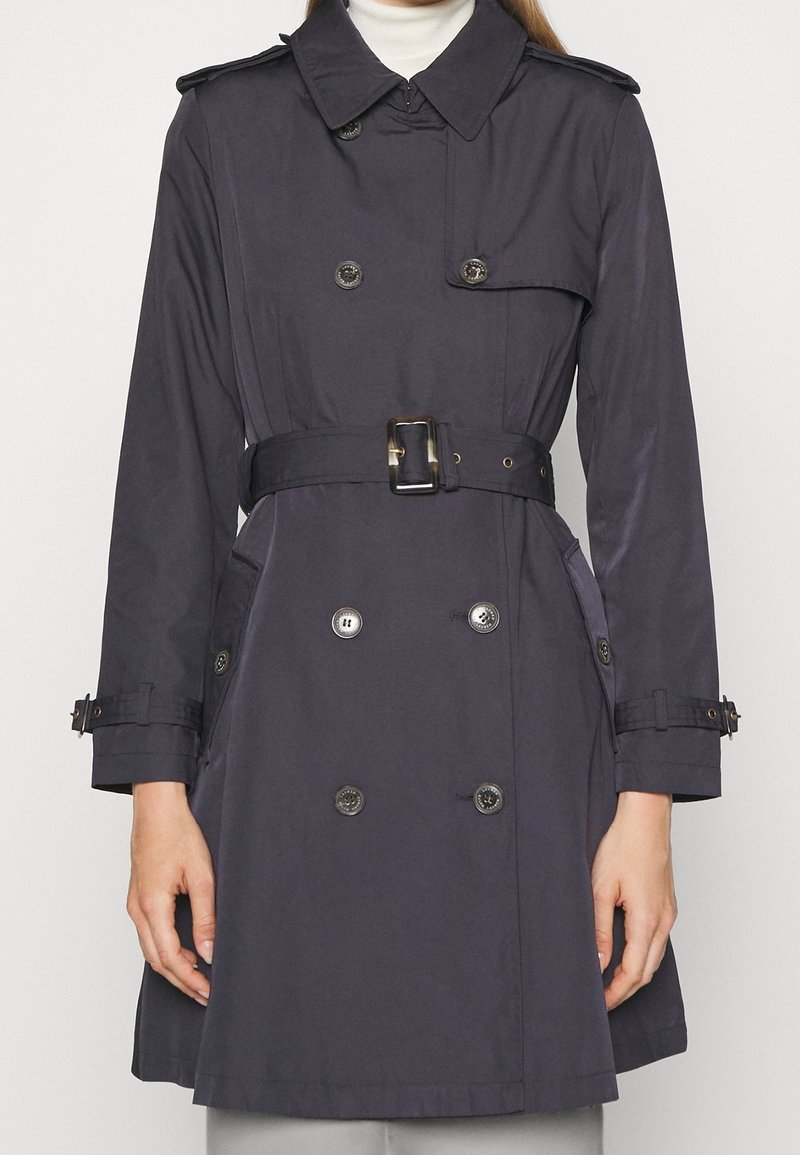 Mörkblå trenchcoat med dubbelknäppt design, stora knappar, justerbart bälte och axelroyaler. Har sidofickor och klassisk krage.