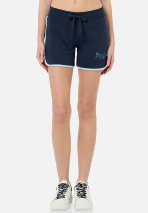 Frau trägt dunkelblaue Shorts mit Kordelzug und hellem Saum sowie weiße Turnschuhe mit schwarzen Schnürsenkeln, stehend vor weißem Hintergrund.
