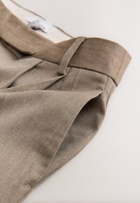 Pantaloni marroni a pieghe con tessuto strutturato e sottili righe a spina di pesce. Presentano una vita piatta e dettagli sartoriali.