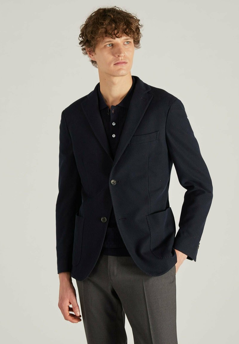 Marineblazer aus strukturiertem Stoff, einreihiges Design, zwei Fronttaschen und mit Revers, kombiniert mit einem dunklen Polo und grauen Hose.