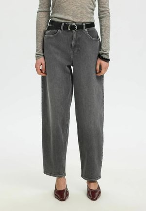Personne portant un jean noir délavé taille haute, un haut gris à manches longues, une ceinture noire et des chaussures pointues rouge foncé, debout avec les mains détendues.