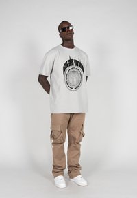 Hellgraues Grafik-T-Shirt mit schwarzem kreisförmigem Design auf dem Rücken, kombiniert mit beigen Cargo-Hosen mit mehreren Taschen und weißen Sneakers.