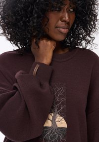 Bruine sweatshirt met een geborduurd boommotief aan de voorkant, met een ronde vorm die wortels en een landschap afbeeldt. Zachte textuur, ontspannen pasvorm.
