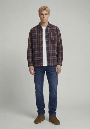 Jeune homme debout portant une chemise à carreaux marron, un t-shirt blanc, un jean bleu et des chaussures marron sur un fond clair uni.
