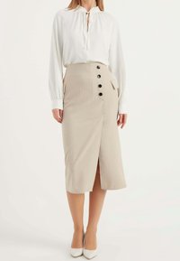 Jupe crayon beige à rayures avec une fente haute et six boutons noirs d'un côté. Associée à un chemisier blanc à manches longues et des escarpins blancs.